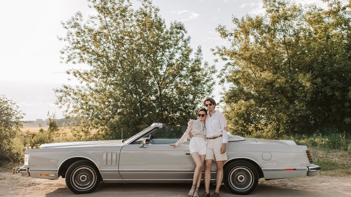 A couple poses beside a vintage convertible on a sunny day in the countryside.